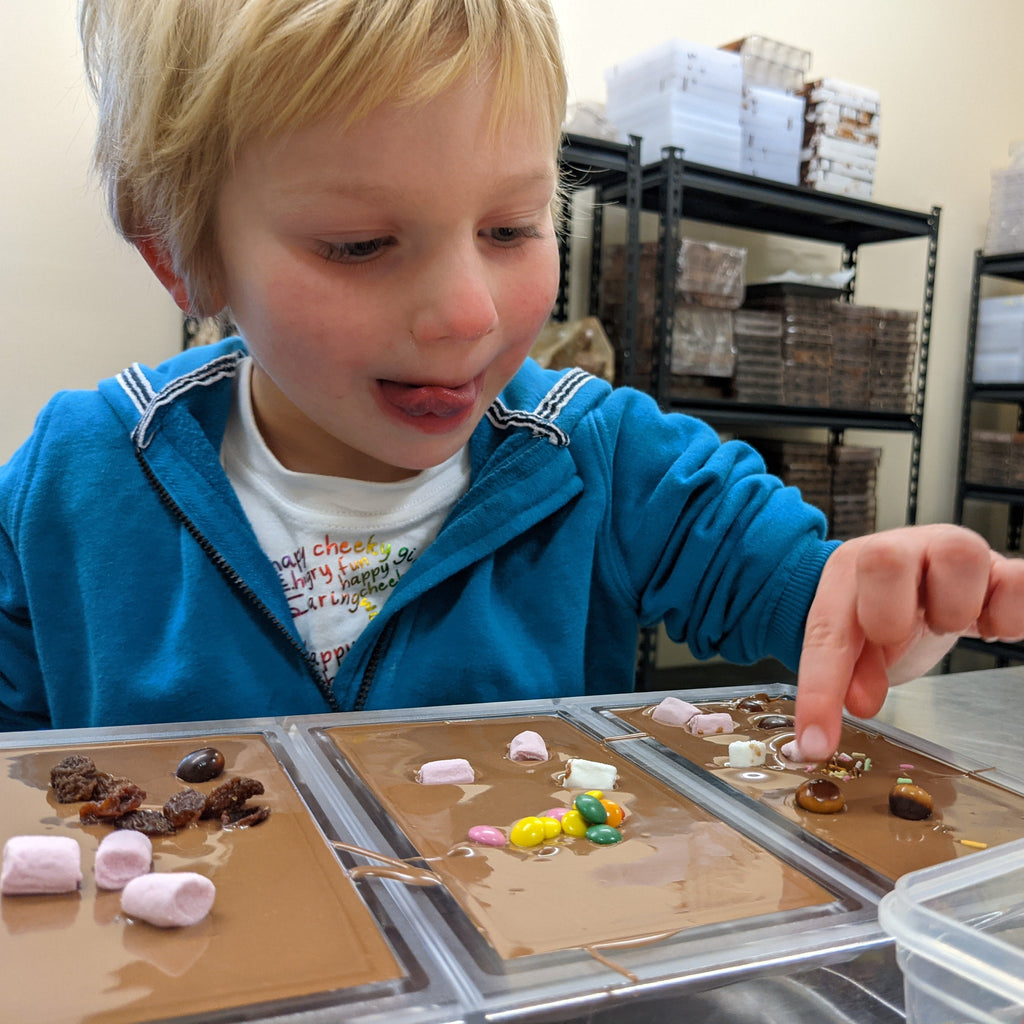 Child in a blue jacket making chocolate bars with shelves in the background.